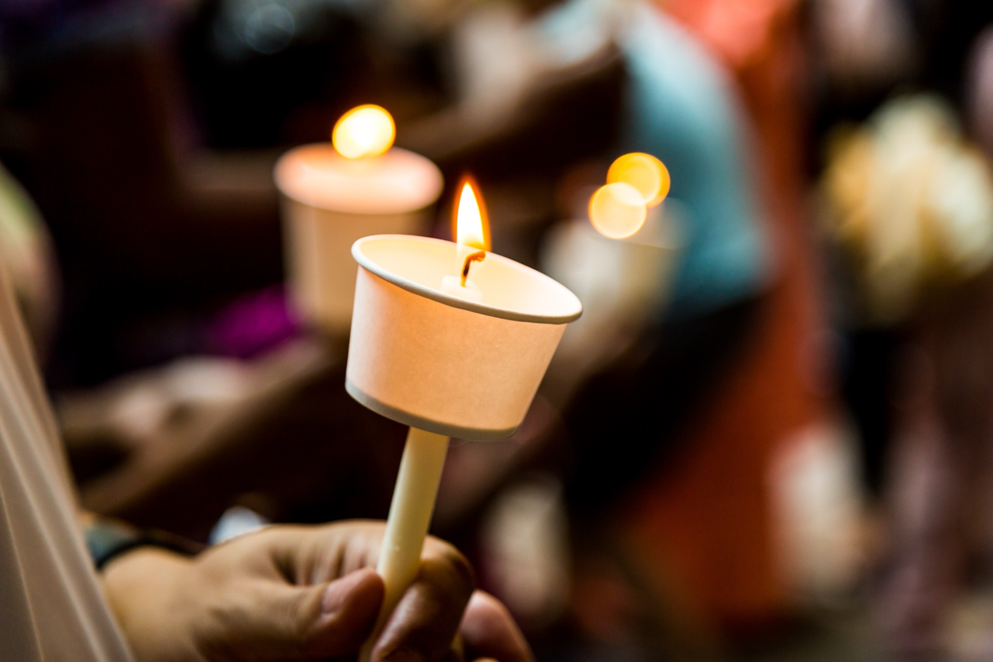 A hand holding a lit candle at a vigil, with more candles glowing softly in the background